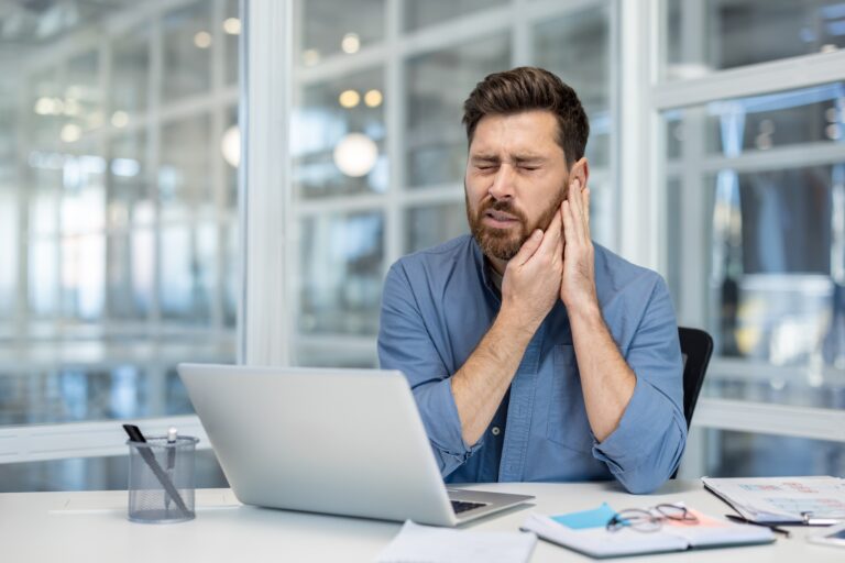 A businessman at his desk in the office is suffering from jaw pain and holding his face, TMJ related pain