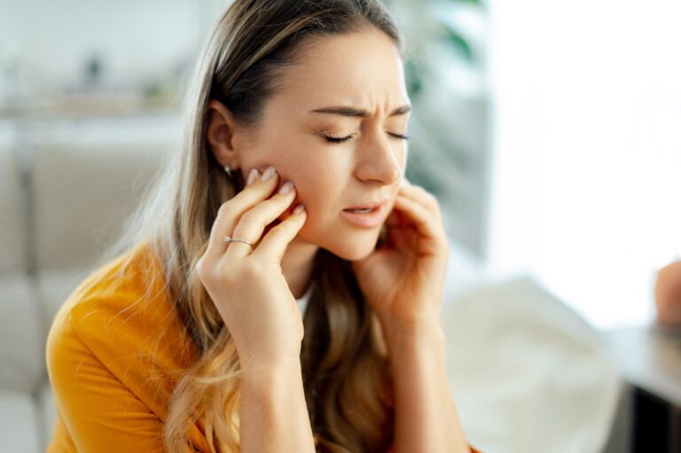 young woman in a yellow shirt sits on a couch at home with eyes closed, pressing her jaw with both hands and wincing, indicating TMJ pain