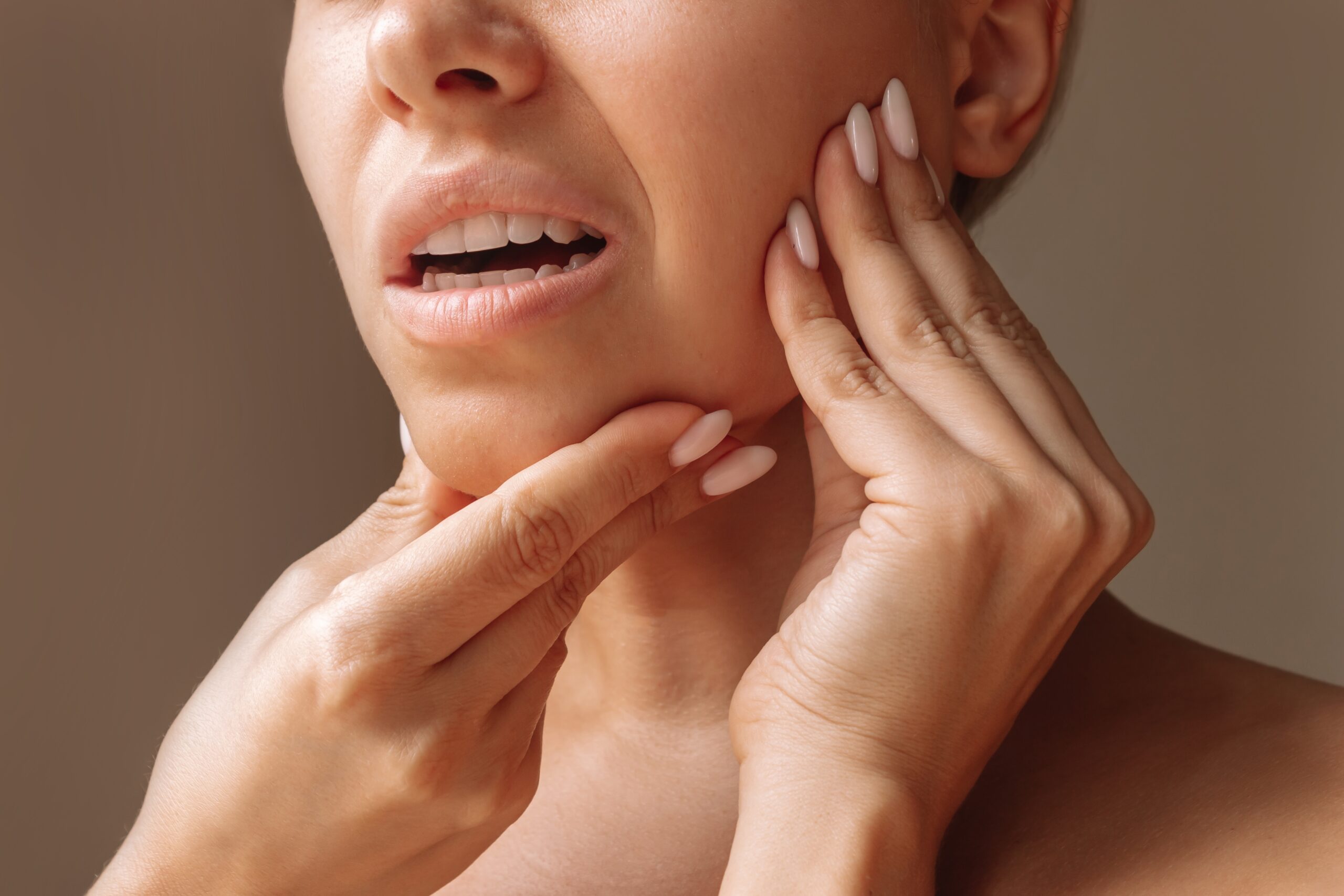 Cropped shot of young woman suffering from jaw pain holding her chin with hands on brown background