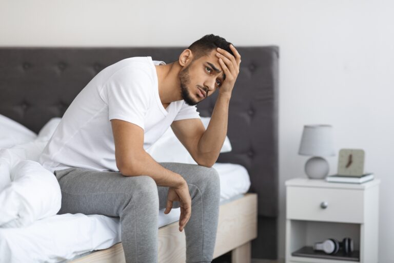 Portrait Of Depressed Young Middle Eastern Man Sitting On Bed In Bedroom, tired with hand on head