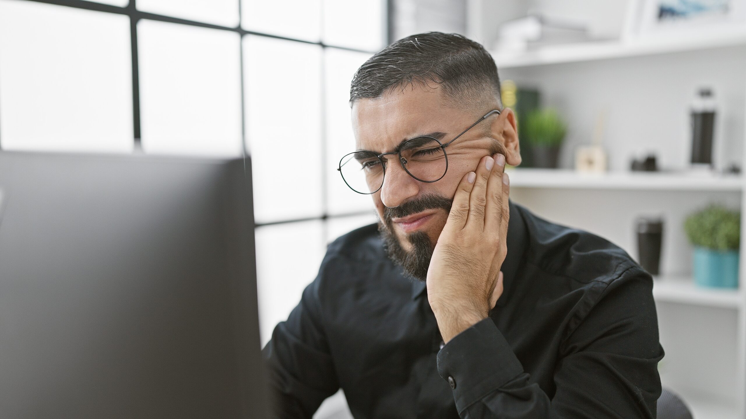young bearded man at desk rubbing jaw, TMJ pain