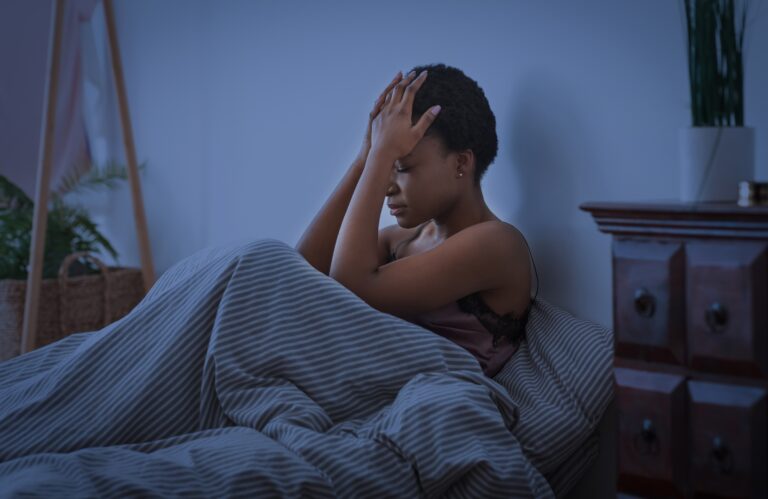 woman sitting up in bed with her hands on head, struggling to sleep