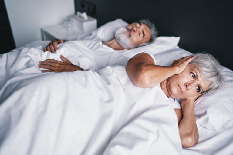 An elderly couple in bed. A rather elderly woman is distressed as her sleeping husband snores