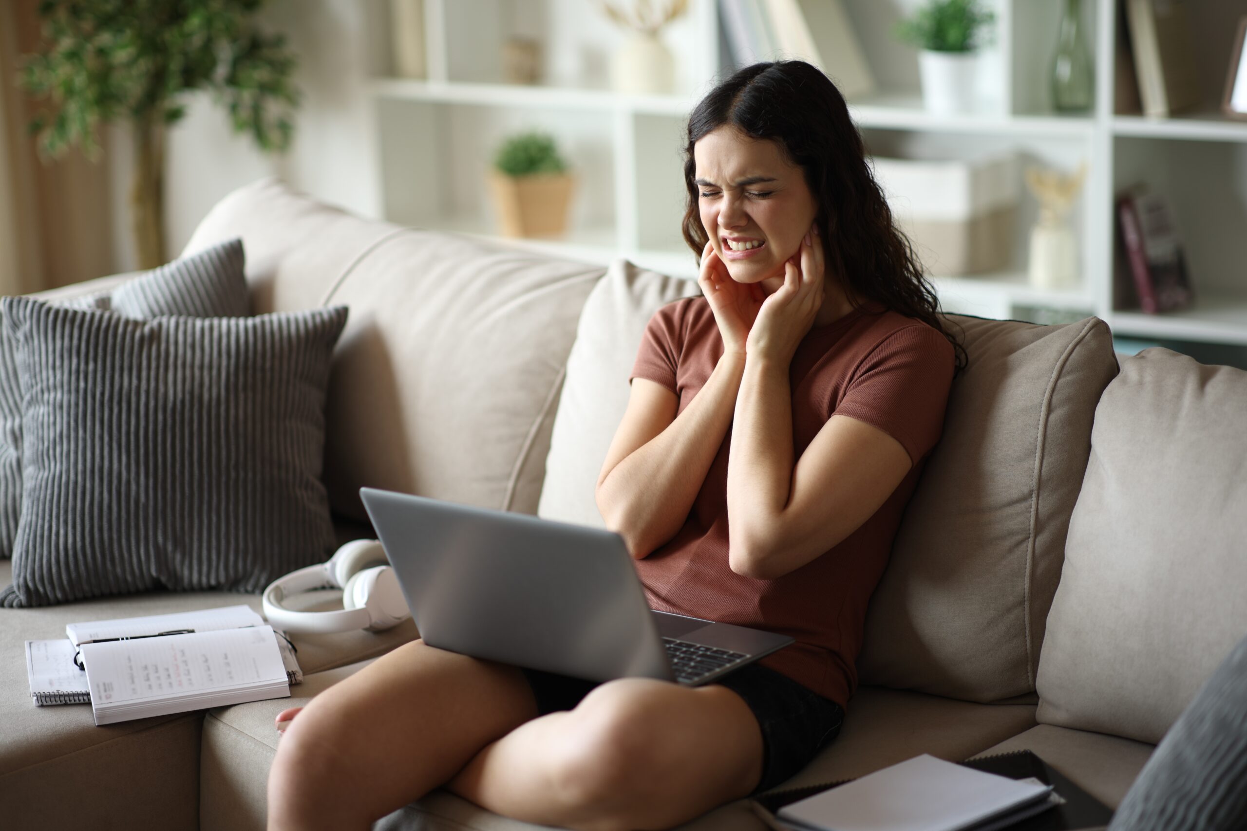 student sitting on couch with laptop, hands on jaw, TMJ pain