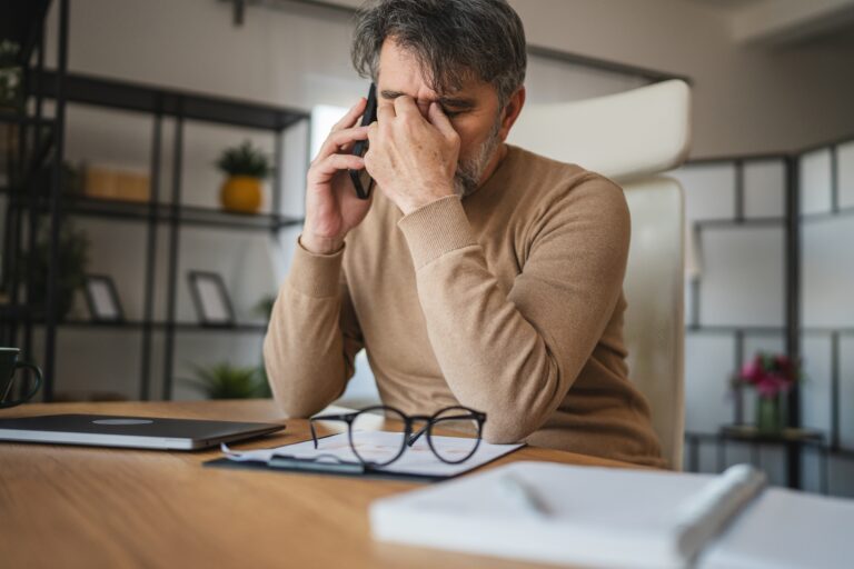 tired adult man works from home and has a conversation about eye strain and pain on his mobile phone